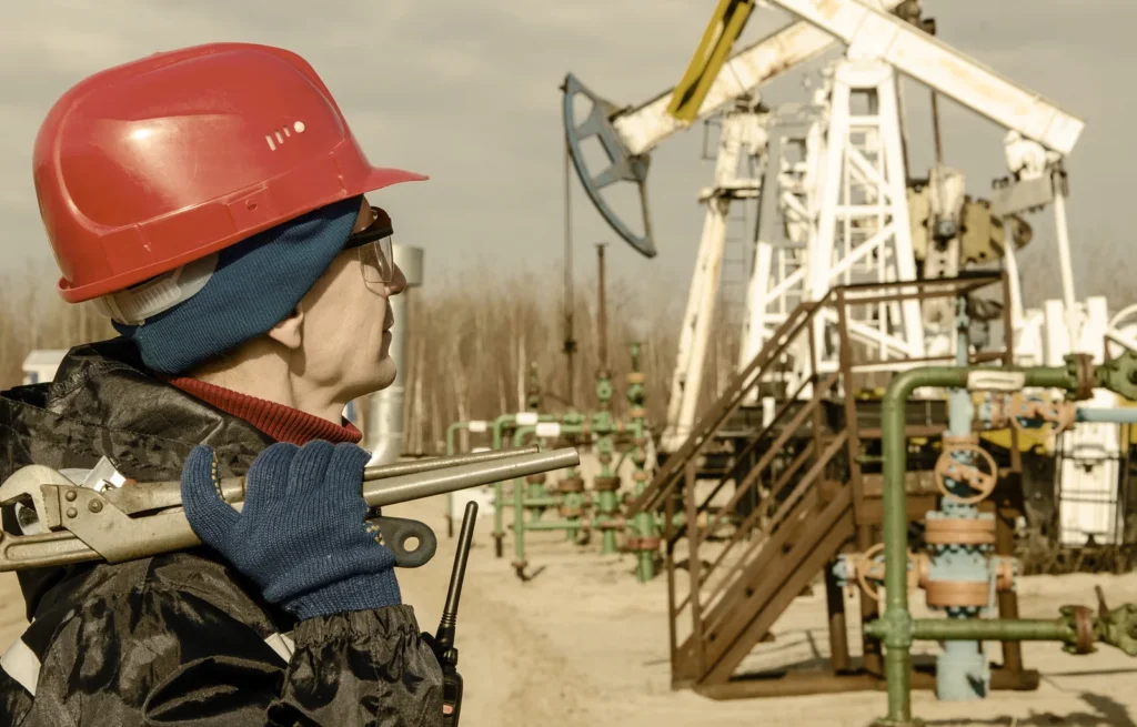 Portrait of man engineer in the oil field wearing red helmet and work clothes holding wrenches in his hand and radio in jacket pocket. Blurry pump jack and wellhead background. Oil and gas concept. Toned.